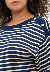 Marineblå og beige stripet topp med rund hals, som har gullknapper på skuldrene og et mykt, teksturert stoff.
