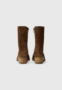 Bottes en daim marron jusqu'aux genoux avec un léger talon, dotées de fermetures éclair à l'arrière et d'un talon en bois texturé.