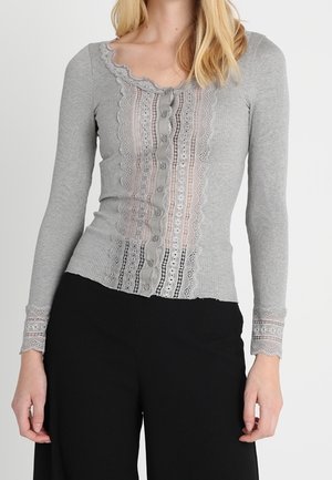 Femme portant un cardigan gris ajusté à manches longues boutonné avec bordure en dentelle et un pantalon noir à jambes larges sur un fond uni.