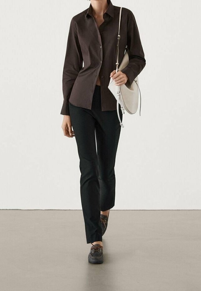 Femme portant une chemise marron foncé boutonnée, un pantalon noir, des chaussures bateau foncées et portant un sac à bandoulière blanc, marchant vers l'avant.