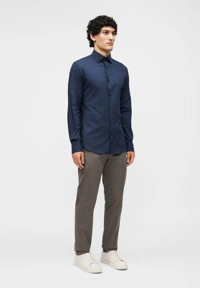 Άνδρας όρθιος, φορώντας μπλε σκούρο πουκάμισο με κουμπιά, γκρι παντελόνι και λευκά αθλητικά, κοιτάζει ελαφρώς αριστερά μπροστά σε απλό φόντο.