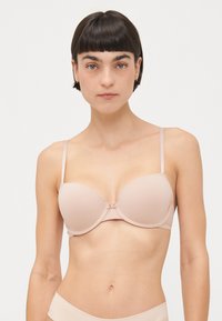 Lys beige formet bh med justerbare stropper, spilerstøtte og en delikat sløyfe i midten. Glatt stofftekstur.