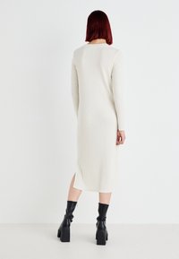 Vestido longo, canelado, de creme, com uma alta fenda no lado esquerdo, apresentando mangas longas e um decote redondo, combinado com botas pretas de salto.