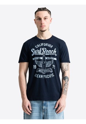 Hombre con el pelo trenzado y tatuajes lleva una camiseta gráfica azul marino "California Surf Beach" y vaqueros azul claro, de pie contra un fondo blanco.