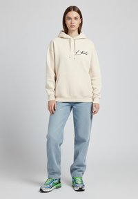Felpa crema con cappuccio e coulisse, con tasca frontale, caratterizzata da un logo nero. Indossata con jeans azzurri chiari e sneakers multicolori.