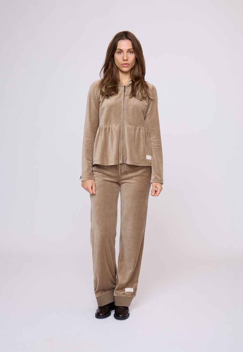 Zamatový dvojdielny outfit na voľný čas v taupe farbe, pozostávajúci z topu s peplum strihom na zips a nohavíc s širokými nohavicami a rebrovanými manžetami. Subtilný detail loga na látke.
