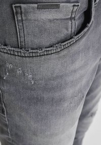Jeans de denim gris con un diseño desgastado, con bordes de bolsillo deshilachados y una etiqueta de marca en relieve en el bolsillo trasero.