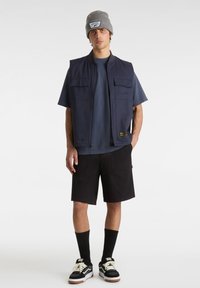 Marinblå ärmlös väst med framfickor över en mörkblå t-shirt, kombinerad med svarta shorts och svarta sneakers med vita detaljer samt svarta strumpor.