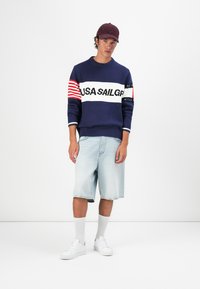 Blue knit sweater with a white band reading "USA SAILGP" and red stripes, paired with light denim shorts and white sneakers. Maroon cap worn.