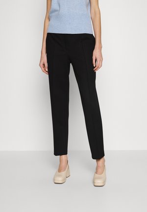 Vrouw draagt zwarte broek met rechte pijpen, lichtblauw mouwloos topje en beige plateauhakken, staand tegen een effen lichte achtergrond.