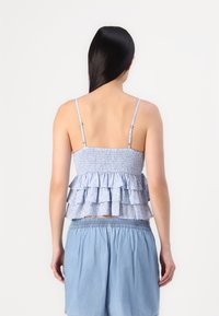 Svetlomodrý volánkový top s nastavitelnými tenkými ramienkami a smúdenou prednou časťou; vrstvený dizajn s kvetinovým vzorom; v kombinácii s modrými šortkami.