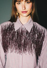 Camicia a righe con un design testurizzato a frange in perline di un bordeaux scuro nella zona del petto; include un colletto classico e una chiusura con bottoni sul davanti.