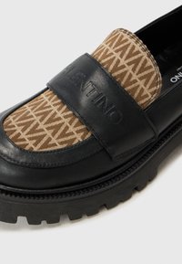 Black leather slip-on shoe with brown patterned textile panel, featuring a wide black strap embossed with "VALENTINO" and a chunky rubber sole.