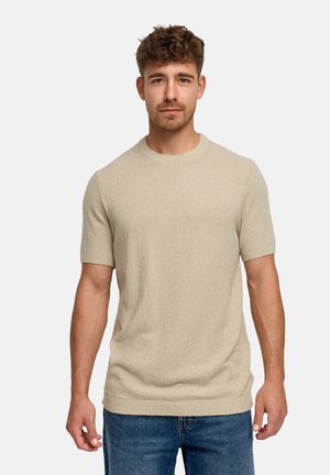 Jonge man met kort krullend haar, die een beige korte mouw gebreide trui en blauwe spijkerbroek draagt, kijkt recht vooruit tegen een effen achtergrond.