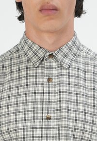 Camisa de cuadros con patrón de checks en beige y negro. Con un collar de punta y botones marrones en una tela texturizada.