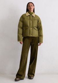 Olivgrüne Daunenjacke mit beigem Kragen, Druckknopfverschluss und zwei Fronttaschen. Kombiniert mit weiten Cordhosen in passender Farbe.
