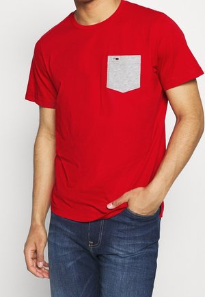 Red cotton t-shirt with a gray pocket. Short sleeves, crew neck design. Pocket features a small logo accent. Worn with dark denim jeans.