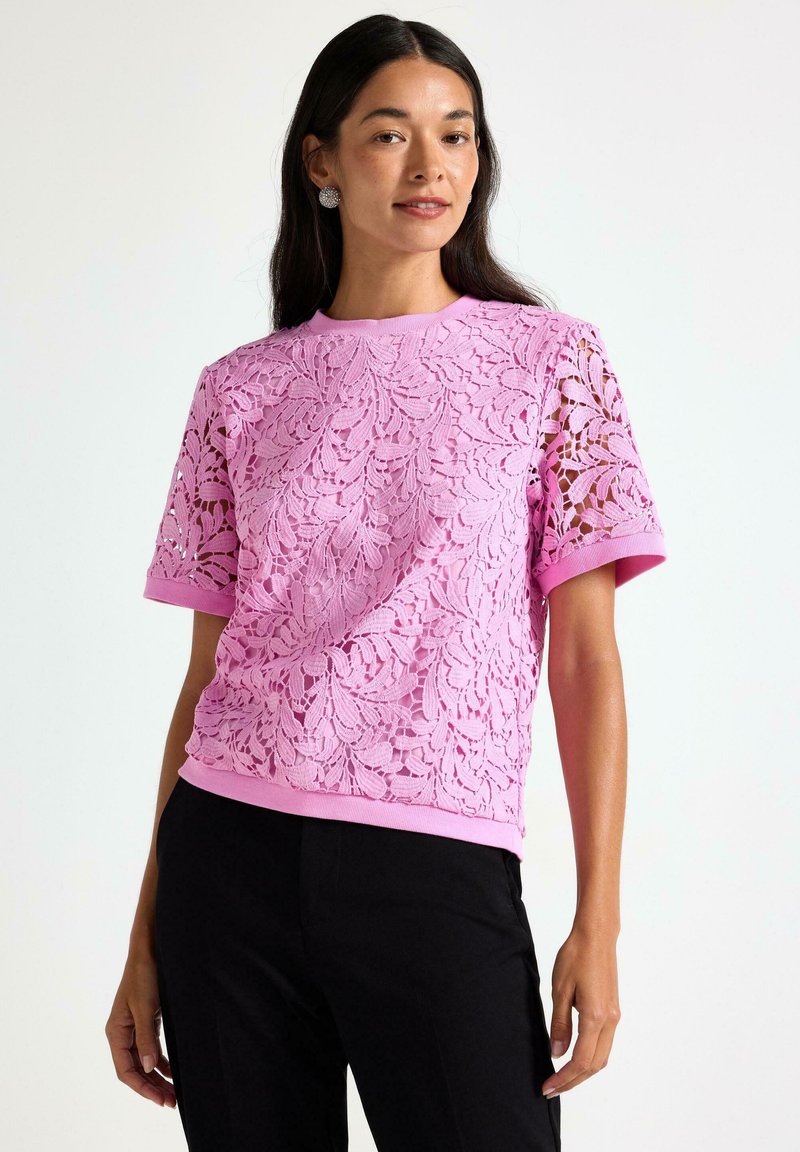 Nainen, joka on pukeutunut vaaleanpunaisiin kukkakuvioisiin pitsitoppeihin lyhyillä hihoilla, jotka on yhdistetty mustiin housuihin, seisoo yksinkertaisen taustan edessä.