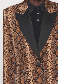 Blazer à motif peau de serpent marron et noir avec un revers en satin noir, une fermeture à un bouton, et une chemise noire à col côtelé en dessous.