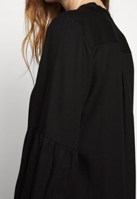 Schwarzes Langarmoberteil mit leicht gerafftem Bund, featuring einer glatten Textur, rundem Ausschnitt und dezenten Falten entlang des Rückens.