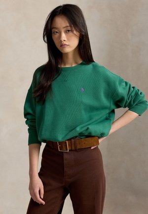 Jeune femme aux longs cheveux foncés porte un sweat vert avec un logo violet, un pantalon marron et une large ceinture marron, posant devant un fond neutre.