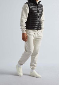 Un gilet matelassé noir avec fermeture éclair sur un sweat à capuche gris clair et un jogging crème, complété par des baskets blanches et des poignets côtelés.