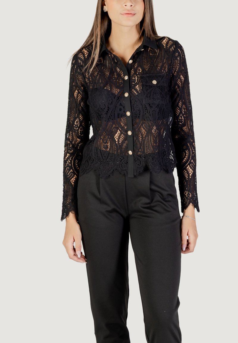 Camicia in pizzo nero con maniche lunghe, chiusura frontale con bottoni, motivi intricati e bordi ondulati, abbinata a pantaloni neri.