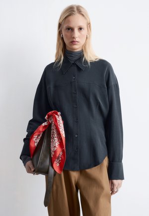 Kobieta ubrana w czarną koszulę zapinaną na guziki i brązowe spodnie, trzymająca ciemną torebkę związaną czerwonym kwiecistym szalikiem, na jednolitym tle.