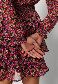 Blommig klänning i svart genomskinligt tyg med rosa och gula blommor, elastisk smockad midja, volangärmar och lager av fåll.