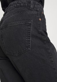 Primo piano di una persona che indossa jeans in denim neri a vita alta, mostrando la tasca posteriore e i rivetti di rame sulla cucitura laterale.