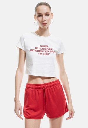 T-shirt blanc raccourci avec du texte rouge qui dit "OUPS SI J'AVAIS L'AIR INTÉRESSÉ, MON BÉBÉ, CE N'EST PAS LE CAS." Associé à des shorts rouges à cordon de serrage. Tissu lisse, style décontracté.