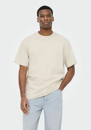 Mand iført en beige, ribstrikket kortærmet skjorte og lyseblå jeans, stående med den ene hånd i lommen mod en ensfarvet baggrund.