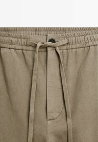 Béžové šortky vyrobené z měkké tkaniny s elastickým pasem, stahovací šňůrkou a jednou přední kapsou. Obsahují detail s knoflíkem.