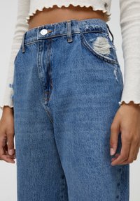 Vaaleansiniset korkeavyötäröiset farkut, joissa on kulutetut yksityiskohdat ja repeämät etutaskuissa, sekä oranssit ompeleet.