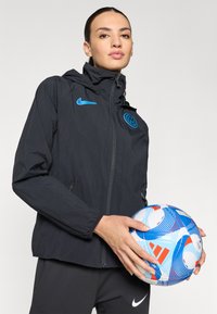 Black lightweight jacket with a hood, featuring a blue Nike logo and a round team emblem. Model holds a blue and white soccer ball.