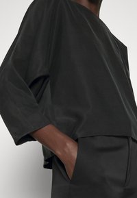 Haut noir ample avec un décolleté large et des manches trois-quarts, fabriqué à partir d'un tissu léger et lisse. Présente un design simple.