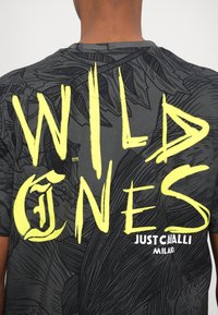 T-shirt graphique noir avec un texte jaune en caractères gras "WILD ONES" et "JUST CAVALLI MILANO." Le tissu présente un motif texturé de feuilles.