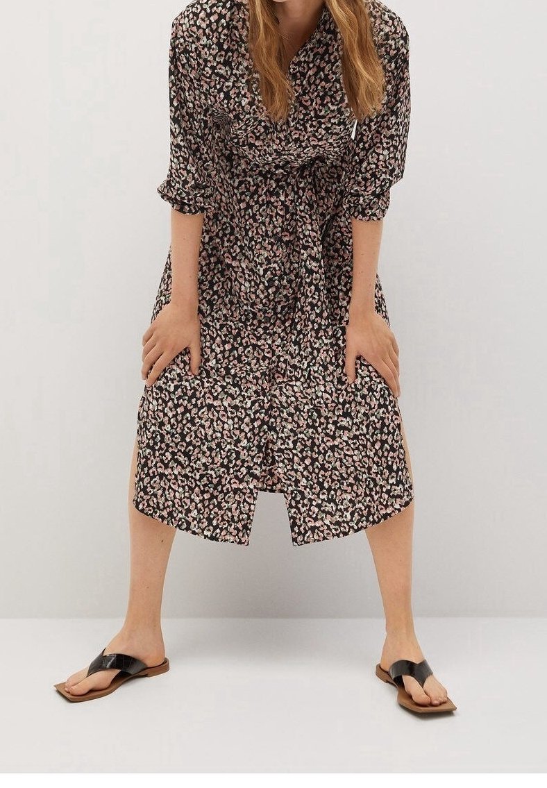 Femme en robe noire mi-longue à motif floral rose, penchée en avant avec les mains sur les genoux, portant des sandales noires sur un sol blanc.