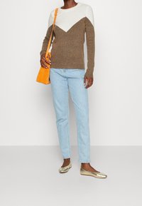 Brązowo-kremowy sweter w zygzakowy wzór, jasnoniebieskie jeansy o prostych nogawkach, metaliczne złote baleriny oraz pomarańczowa torba na ramię.