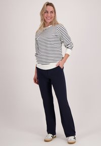 Lachende vrouw staat met één hand in haar zak, gekleed in een wit met zwart gestreept shirt met lange mouwen, zwarte broek en witte sneakers.