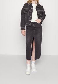 Černá džínová bunda s knoflíkovým zapínáním a kapsami na hrudi, doplněná dlouhou černou džínovou sukní s předním rozparkem, nošená s bílými teniskami.