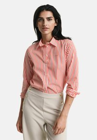 Camicia a righe rosse e bianche con colletto, dotata di maniche arrotolate e discreto logo. Abbinata a pantaloni beige a vita alta.