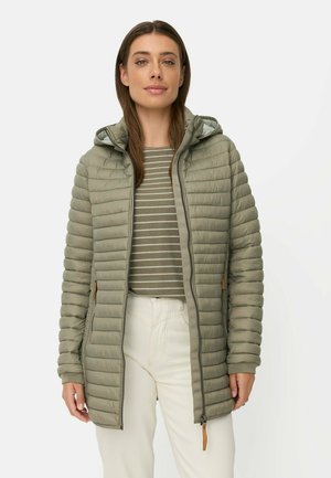 Frau trägt eine hellolivgrüne gesteppte Jacke mit Kapuze, ein gestreiftes grünes und beiges Shirt und weiße Hosen vor einfachem Hintergrund.