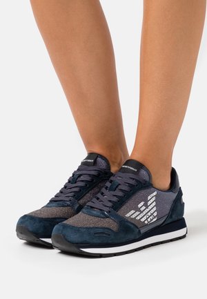 Pieds portant des baskets lacées bleu foncé et gris avec des bandes blanches du logo et des semelles noires et blanches sur fond blanc.