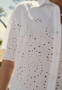 Camicia bianca in cotone con motivo tagliato ricamato, dotata di chiusura con bottoni e colletto. Maniche corte con bordi scallop.