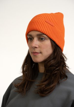 Oranje geribbelde gebreide beanie met een gestructureerde afwerking en een klein logo-label, gedragen door een persoon in een grijze sweatshirt.