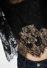 Černá květinová krajka překrývající holou pokožku nad tmavě modrými džínami s viditelným knoflíkem a švy.