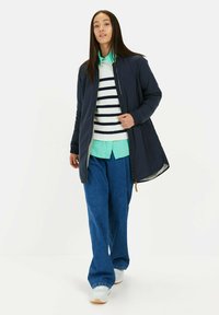 Marineblauwe lange jas met rits aan de voorkant, geribbelde manchetten en zakken, gedragen over een gestreepte trui en een lichtgro shirt, gecombineerd met een wijde broek.