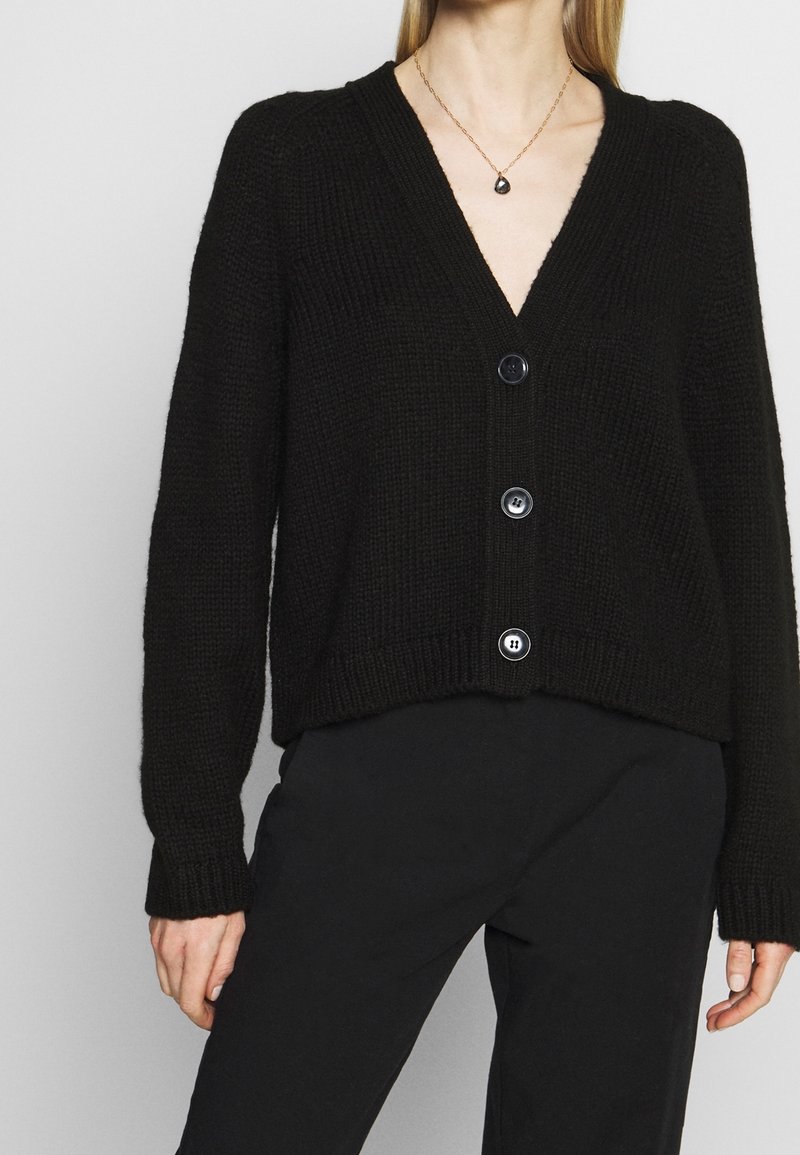 Černý pletený cardigan s výstřihem do V, který má pět velkých knoflíků a žebrované manžety na rukávech, v kombinaci s černými kalhotami. Texturovaná tkanina.
