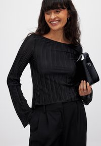 Black, ribbed long-sleeve top with an off-shoulder neckline. Paired with high-waisted black trousers and a small, structured handbag.